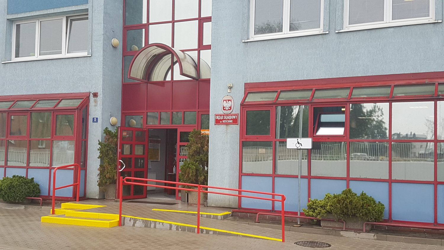 Na zdjęciu budynek w niebieskiej elewacji, wiśniowe obramowania przeszklonego wejścia do budynku