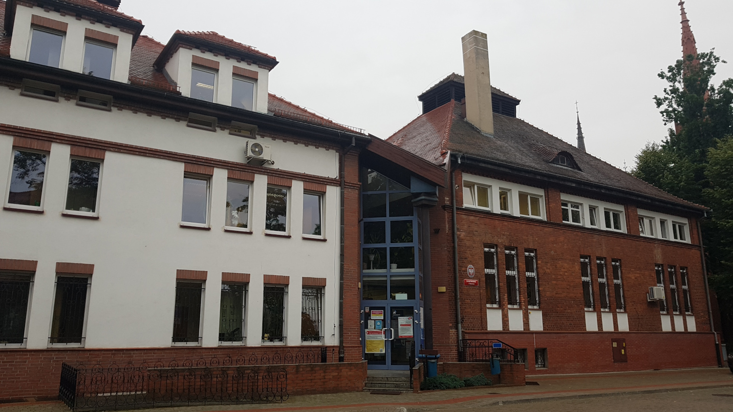 Na zdjęciu budynek w białej elewacji w tym elementy elewacji z cegły, wejście do budynku przeszklone z niebieskimi obramowaniami.