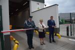 Trzy osoby przecinają biało-czerwoną wstęgę podczas uroczystego otwarcia obiektu; po lewej funkcjonariusz Krajowej Administracji Skarbowej w mundurze, w środku kobieta w jasnej marynarce, po prawej mężczyzna w granatowym garniturze.