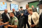 Grupa funkcjonariuszy i gości obserwuje stanowisko pracy funkcjonariusza Służby Celno-Skarbowej w centrum monitoringu, na ekranach w tle widoczne podglądy z kamer.