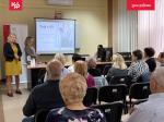 Prowadzące spotkania stoją, obok na ekranie wyświetlana jest prezentacja, na krzesłach siedzą uczestnicy.