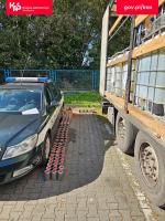 Zdjęcie przedstawia szczegóły kontroli naczepy wypełnionej chemikaliami. Widok z perspektywy boku ciemnozielonego radiowozu Służby Celno-Skarbowej, obok którego, na brukowanym podłożu, ustawiono w równych rzędach kilkadziesiąt małych, metalowych puszek z czerwonymi nakrętkami. Po prawej stronie kadru widoczna jest naczepa ciężarówki z załadowanymi zbiornikami typu IBC (mauzery). Zbiorniki są białe, ponumerowane i opatrzone piktogramami ostrzegawczymi. W tle widać niebieski płot oraz gęste, zielone drzewa. W górnych rogach zdjęcia znajdują się logotypy: lewy: Krajowa Administracja Skarbowa (KAS) oraz prawy: adres portalu gov.pl/kas.