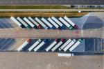 Zdjęcie wykonane z lotu ptaka przedstawia parking dla samochodów ciężarowych. Na wyznaczonych miejscach postojowych zaparkowane są ciągniki siodłowe z naczepami ustawione pod kątem. W górnej części zdjęcia widoczna jest droga z poruszającymi się i zaparkowanymi samochodami osobowymi, a na nawierzchni parkingu znajdują się oznaczenia poziome, w tym znaki zakazu zatrzymywania się.