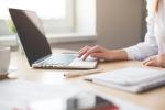 Na zdjęciu widoczny jest otwarty laptop ustawiony na biurku oraz osoba obsługująca go jedną ręką. Na blacie znajdują się także długopis, notes, kubek oraz rozmyte dokumenty na pierwszym planie. W tle widoczne jest okno wpuszczające naturalne światło.
