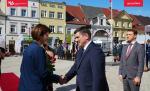 mężczyzna w granatowym garniturze składa gratulacje kobiecie w granatowej marynarce w tle zdjęcia poczty sztandarowe. Za mężczyzną w granatowym garniturze stoi mężczyzna w szary garniturze.