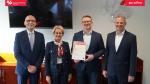 Jedna kobieta  i trzech mężczyzn stoją w jednym rzędzie, uśmiechają się. Wszyscy ubrani w marynarki koloru granatowego. Kobieta ma kolorową apaszkę. Mężczyzna w środku trzyma w rękach dokument w białej ramce.