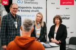 Przy stoisku informacyjnym Krajowej Administracji Skarbowej trzy osoby rozmawiają z uczestnikiem wydarzenia, widocznym tyłem na pierwszym planie. W tle znajdują się roll-upy promujące usługi e-Urzędu Skarbowego oraz Krajowy System e-Faktur (KSeF), z hasłami „Wygodnie. Szybko. W każdej chwili.