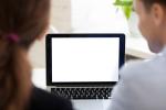 Laptop na biurku oraz jedna dłoń trzyma sprawozdania a druga dotyka klawiatury komputera.