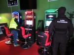 Dwóch funkcjonariuszy w trakcie kontroli automatów w lokalu. Trzy automaty gotowe do gry i trzy fotele dla graczy. Jeden z funkcjonariuszy w kominiarce ubrany w czarny mundur polowy, drugi w zielonej kamizelce